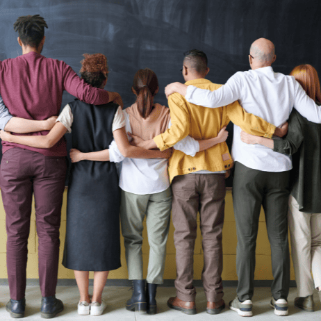 Group of diverse people standing side by side with arms around each other's shoulders, facing a chalkboard.