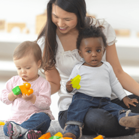A woman playing with two young children on the floor, one child is playing with building blocks while the other holds a toy.